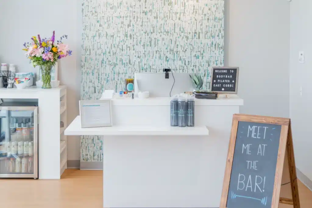 A bright juice bar with a white counter, flower vase, drinks fridge, and a chalkboard sign reading “MEET ME AT THE BAR!”.