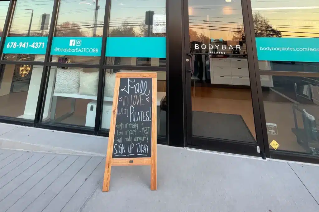 A sidewalk chalkboard sign in front of a Pilates studio with glass doors and teal signage.