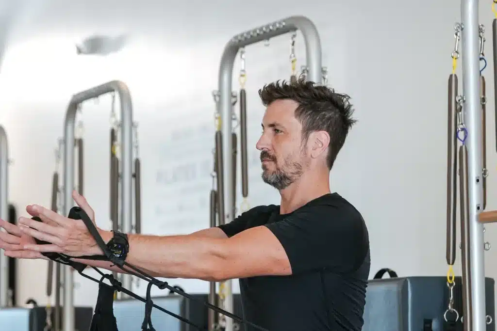 Man doing Pilates exercise with resistance bands in a gym, arms extended forward, wearing a black shirt and watch.