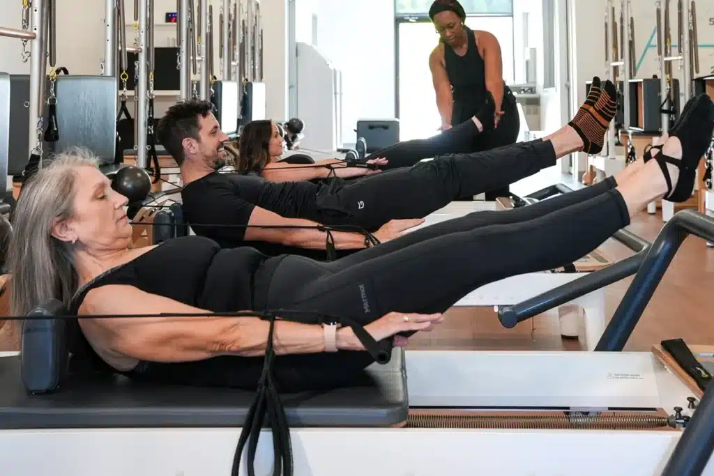 Four people in a Pilates class using reformer machines, stretching their legs, with an instructor assisting.