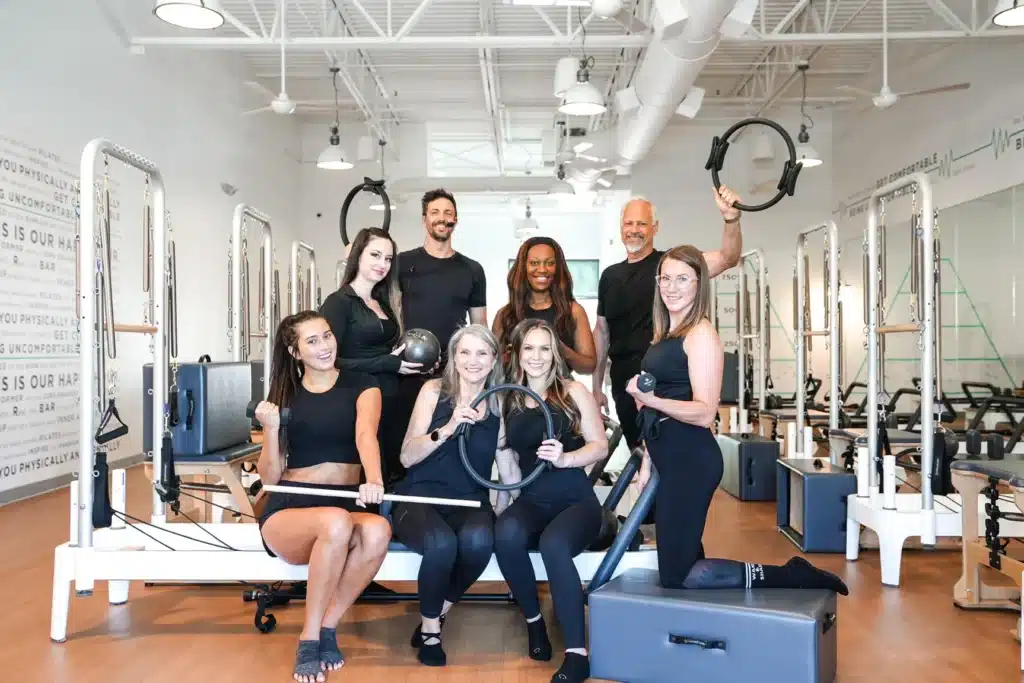 Eight people in a Pilates studio pose with exercise rings and smile at the camera, wearing black athletic clothing.
