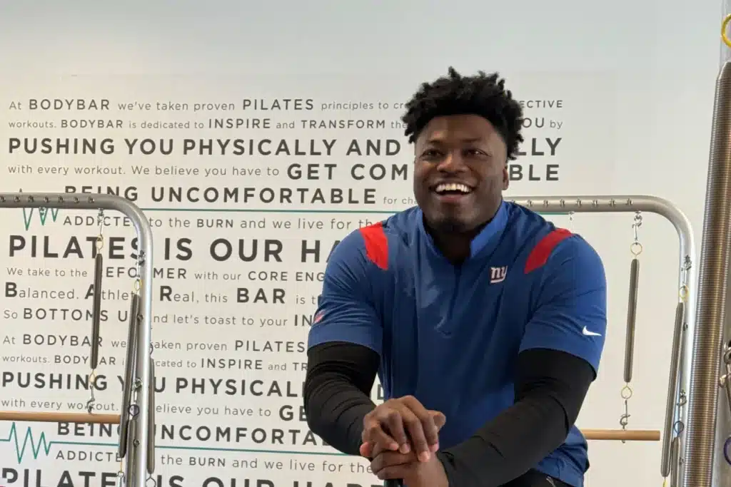 Smiling man in a blue sports shirt sits in a gym with pilates equipment and motivational text on the wall.