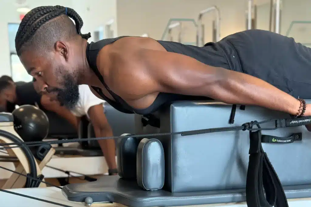 A man works out on a Pilates reformer machine, lying face down and pulling straps with his arms extended back.