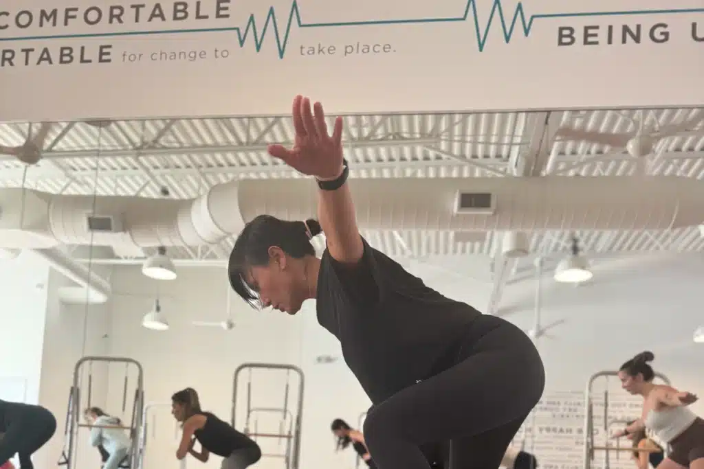 A woman in workout clothes balances on one leg in a fitness studio with others exercising in the background.