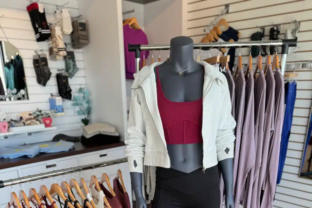 Mannequin in a gymwear store wears a maroon sports bra and gray zip hoodie, with racks of activewear in the background.