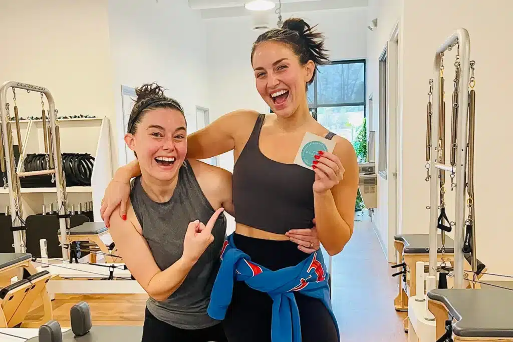 Two women in workout clothes smile in a gym. One points at the other holding a round item and both look happy.