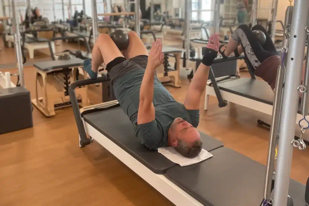 Man lying on a Pilates reformer with feet on a ball, arms raised, exercising in a fitness studio.