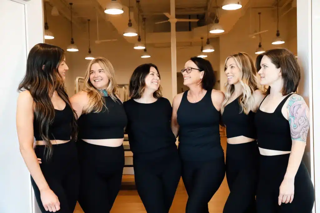 Six women in black workout outfits stand together indoors, smiling and talking to each other in a bright gym studio.