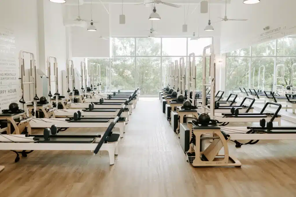 A bright pilates studio with several reformer machines lined up in rows on a wooden floor, large windows in back.