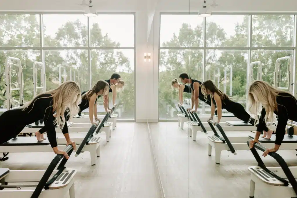 People exercise on reformer machines in a bright fitness studio with large windows and mirrors.
