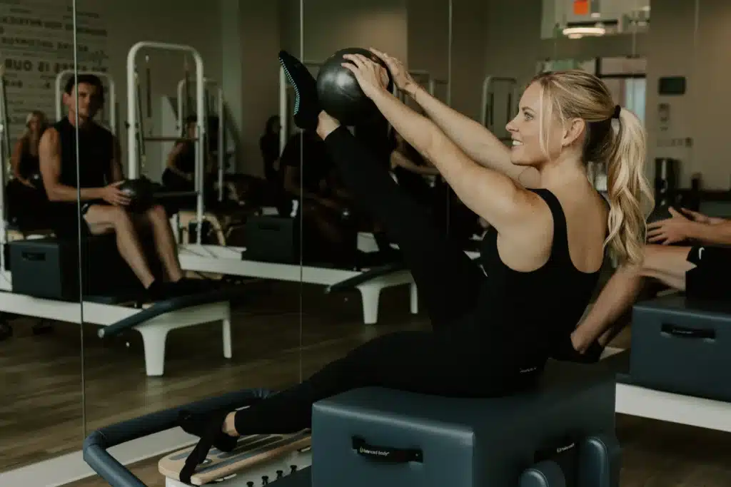 Woman in black workout clothes does Pilates with a ball, lifting leg on equipment in a fitness studio with mirrors.