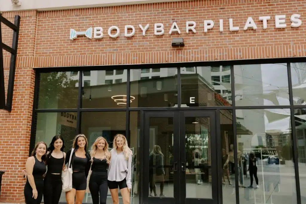 Five women in black athletic wear stand smiling outside a BODYBAR Pilates studio with glass doors and brick exterior.