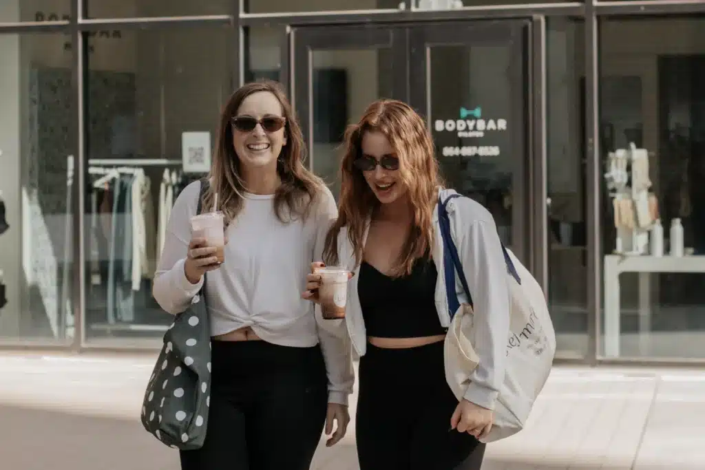 Two women wearing sunglasses and casual outfits walk outside, smiling and holding iced drinks in front of a building.