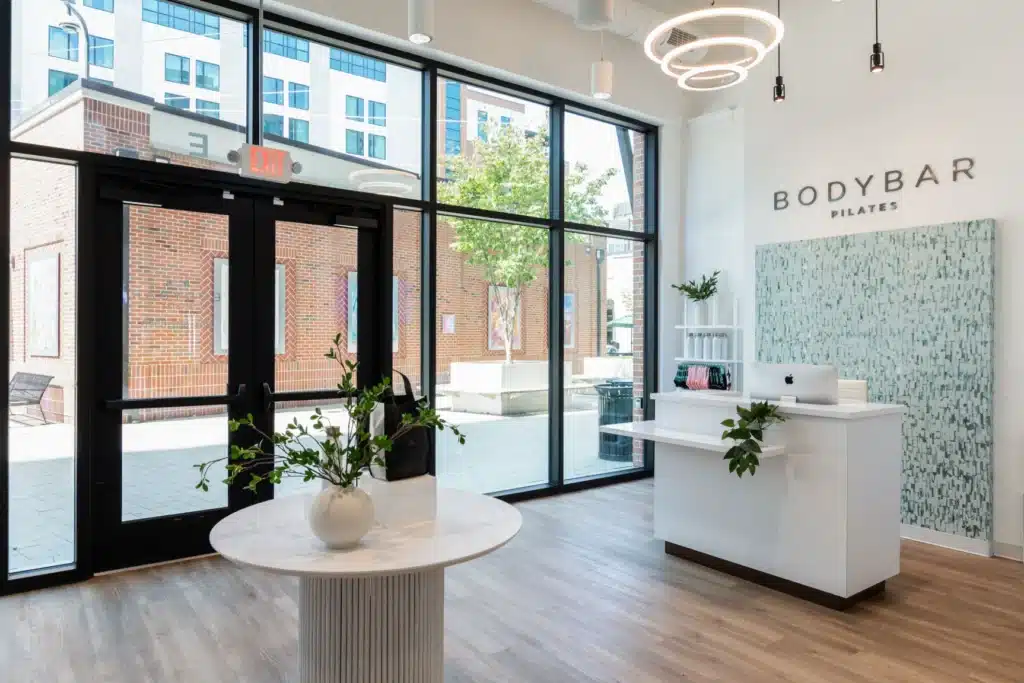 Modern pilates studio lobby with large windows, a round table, and a sleek reception desk with plants.