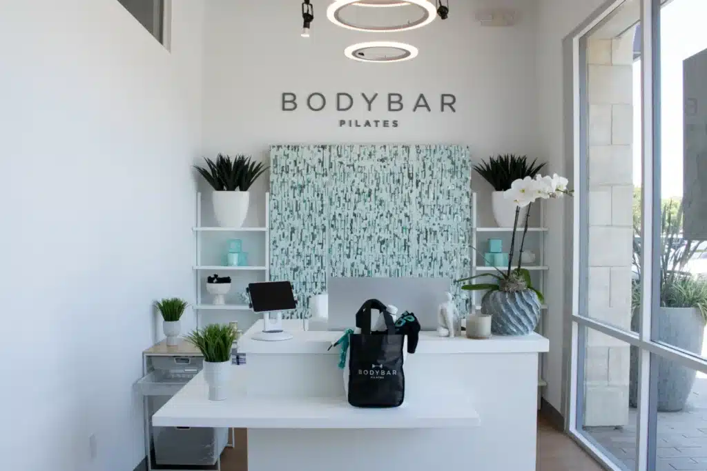 Minimalist Pilates studio front desk with plants, a tablet, and a BODYBAR bag; BODYBAR PILATES sign on back wall.