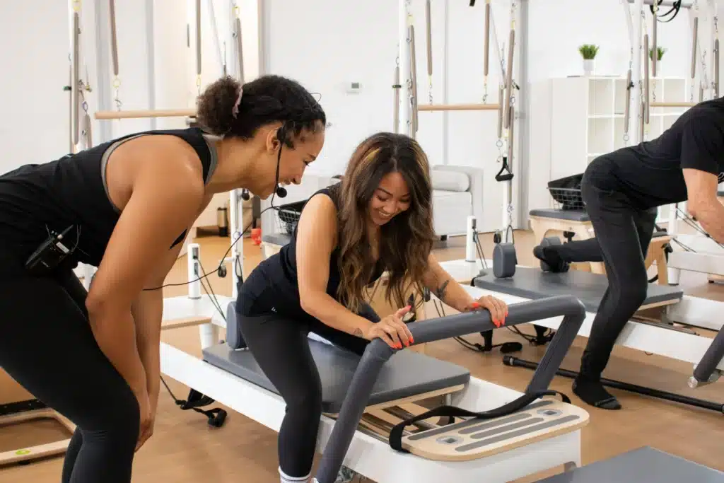 Instructor guides a smiling woman using Pilates equipment in a bright fitness studio. Another person works out nearby.