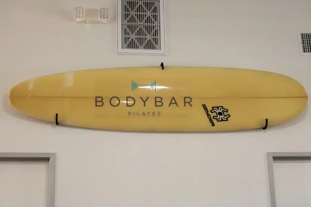 A yellow surfboard with “BODYBAR PILATES” logo is mounted on a white wall above two doorways.