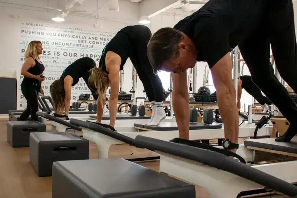 Four people in a pilates studio doing plank exercises on reformer machines with an instructor watching.
