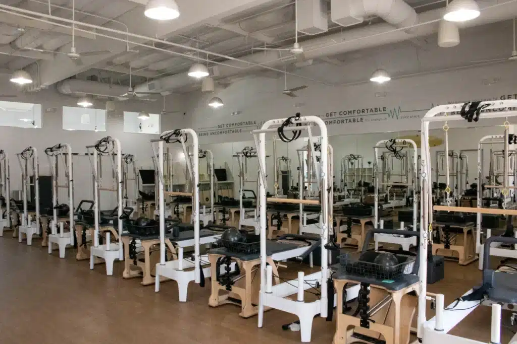 A bright Pilates studio with several reformer machines arranged in rows on a wooden floor.