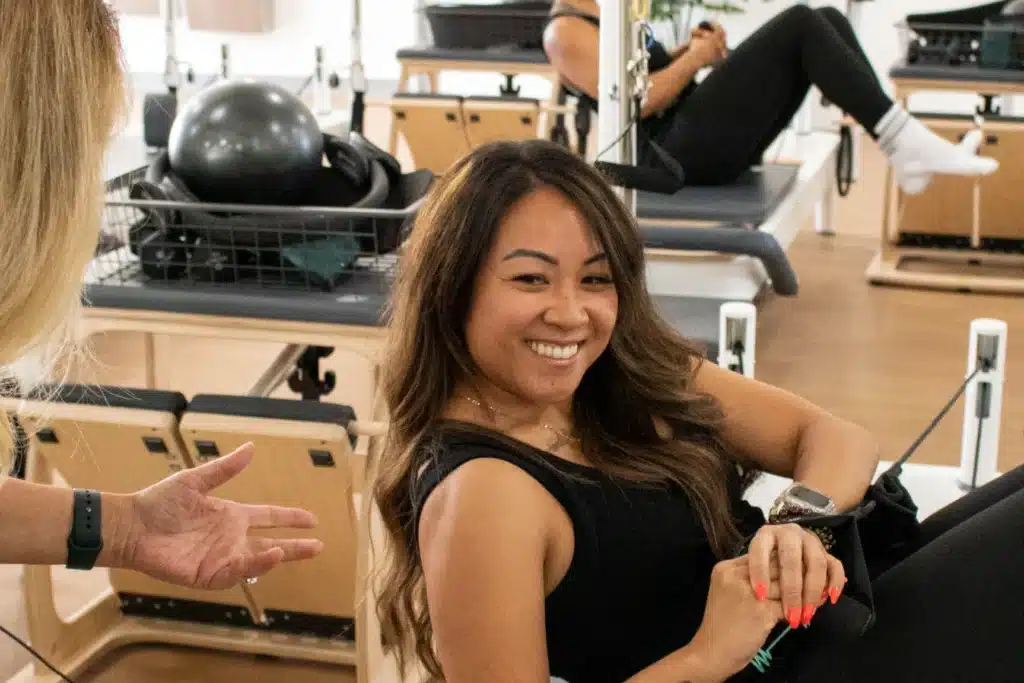 Woman smiling while exercising on Pilates equipment in a gym, with another person’s hand gesturing nearby.