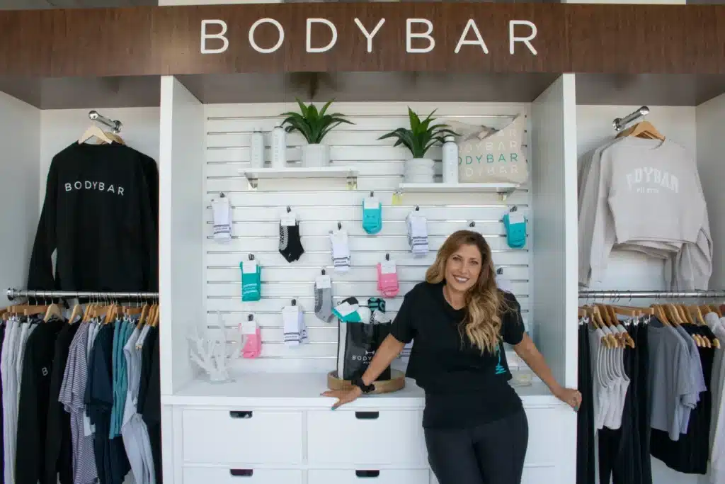 Woman smiling and standing in a BODYBAR store with clothes, socks, and plants displayed on shelves behind her.