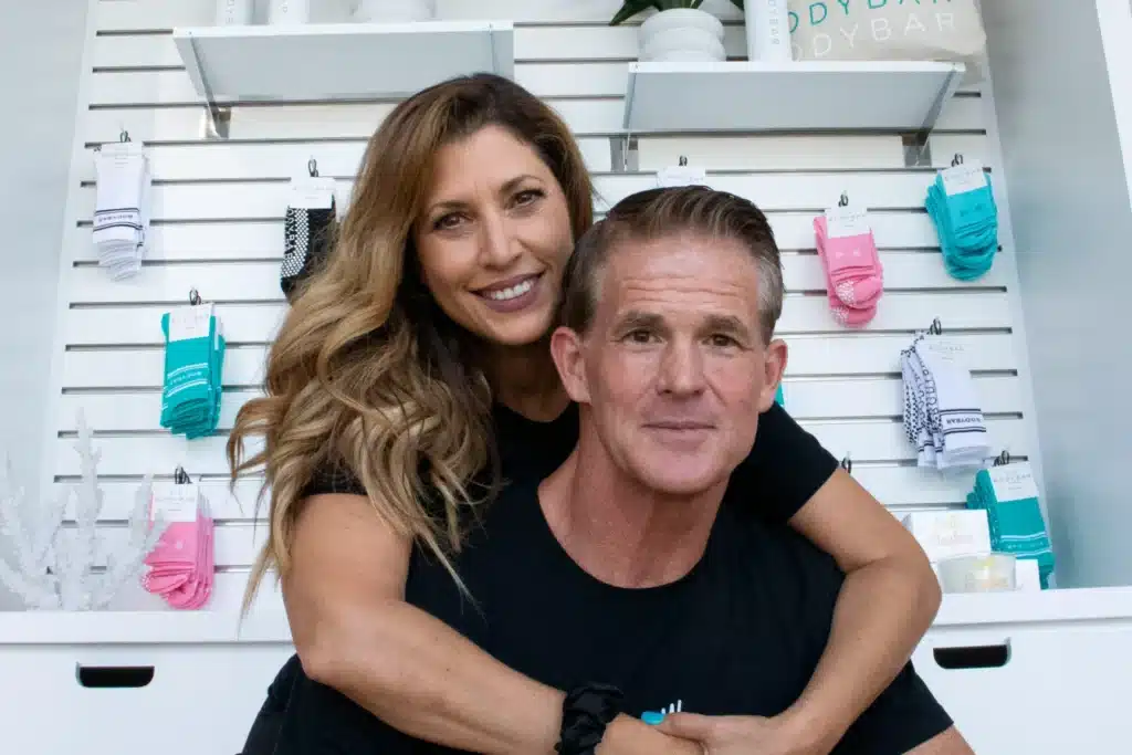 A woman hugs a man from behind as they both smile, standing in front of a display of colorful packaged items.