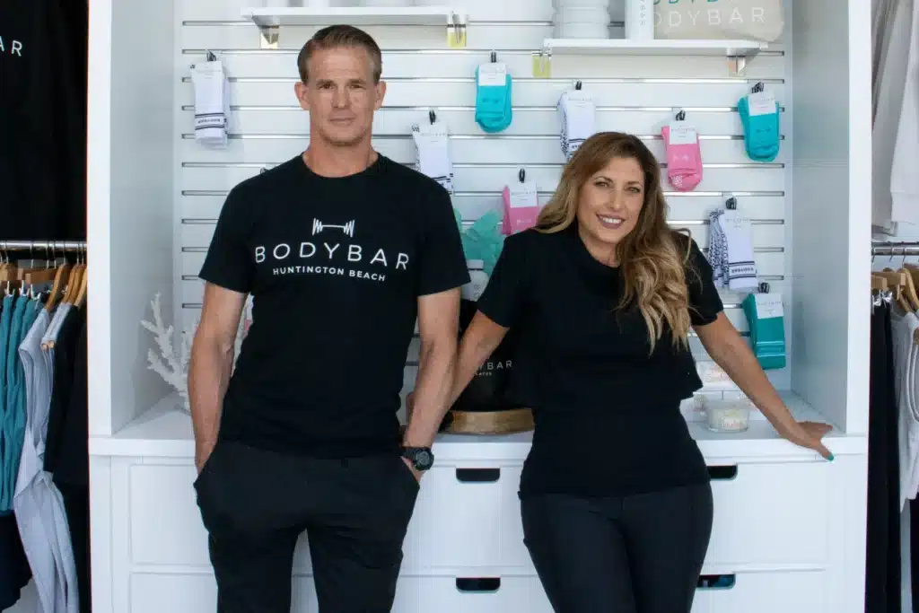 Two people in black BODYBAR shirts stand and smile in a fitness retail store with socks and clothes on display.