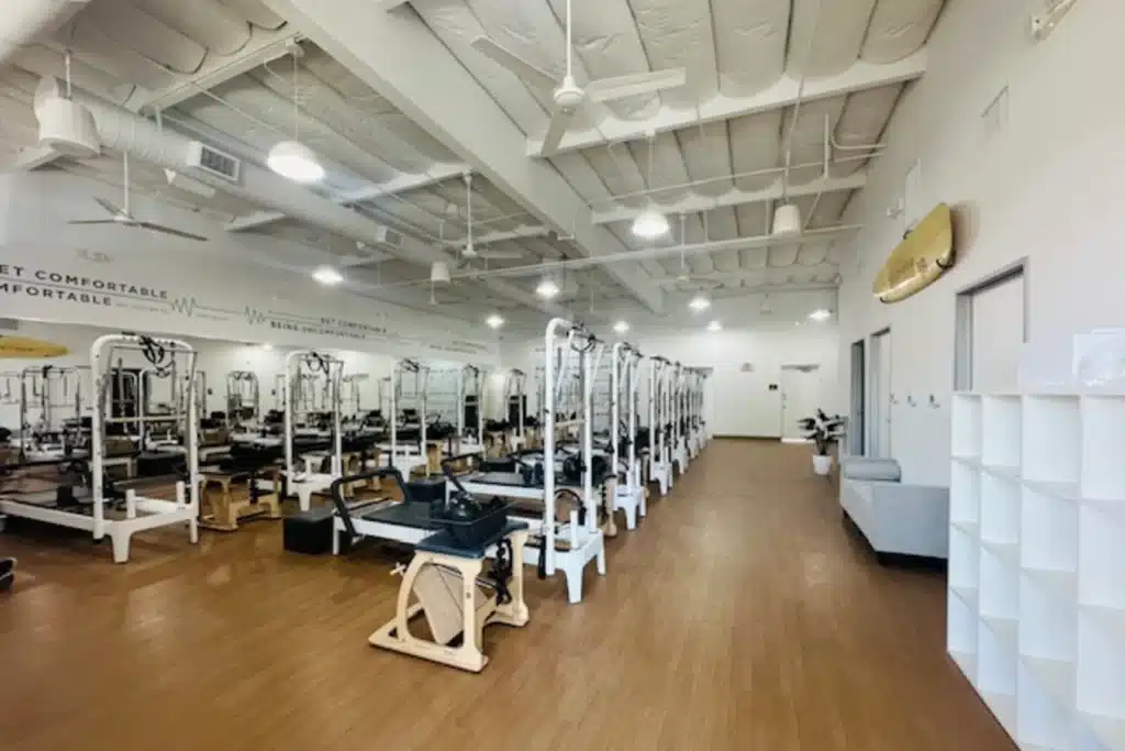 A spacious Pilates studio with reformer machines, white walls, wood flooring, and ceiling fans.