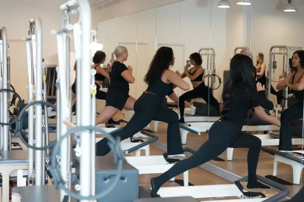 Five women exercise on reformer machines in a fitness studio, performing lunges while facing a mirror.