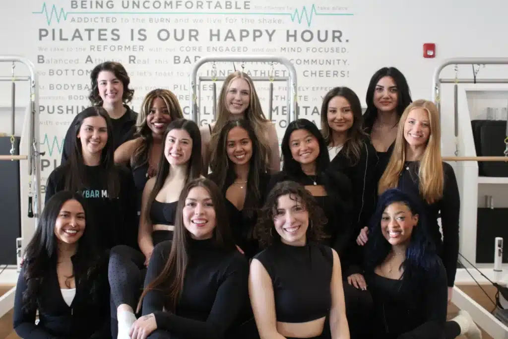 A group of fifteen women in workout clothing pose and smile together in a Pilates studio.