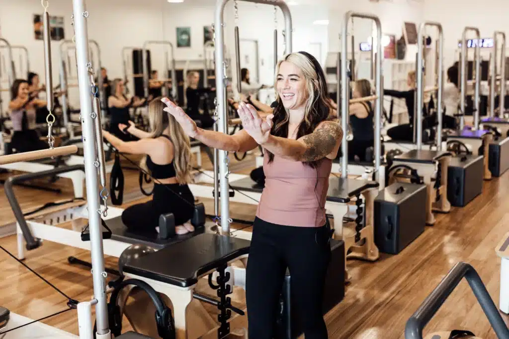 Woman leading a Pilates class, smiling with arms outstretched, in a studio with reformer machines and other participants.