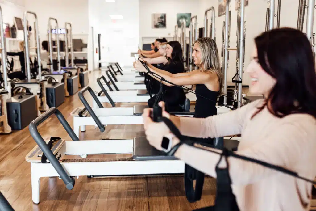 People exercising on Pilates reformer machines in a bright studio, smiling and pulling resistance straps.