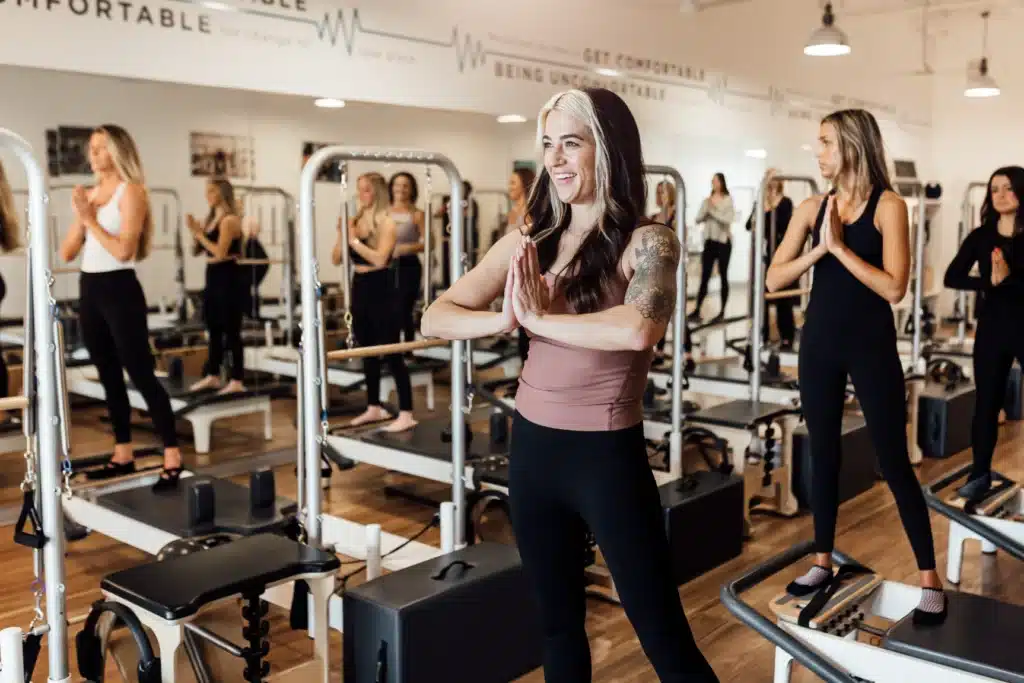 Women in a fitness studio practice a standing pose on Pilates reformer machines, smiling and focused.