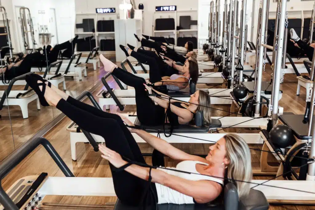People exercising on Pilates reformer machines in a fitness studio, stretching their legs while holding resistance straps.