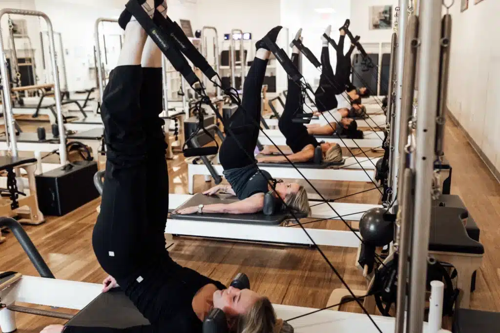 Five people doing Pilates on reformer machines in a bright studio, all lifting their legs in the air during exercise.