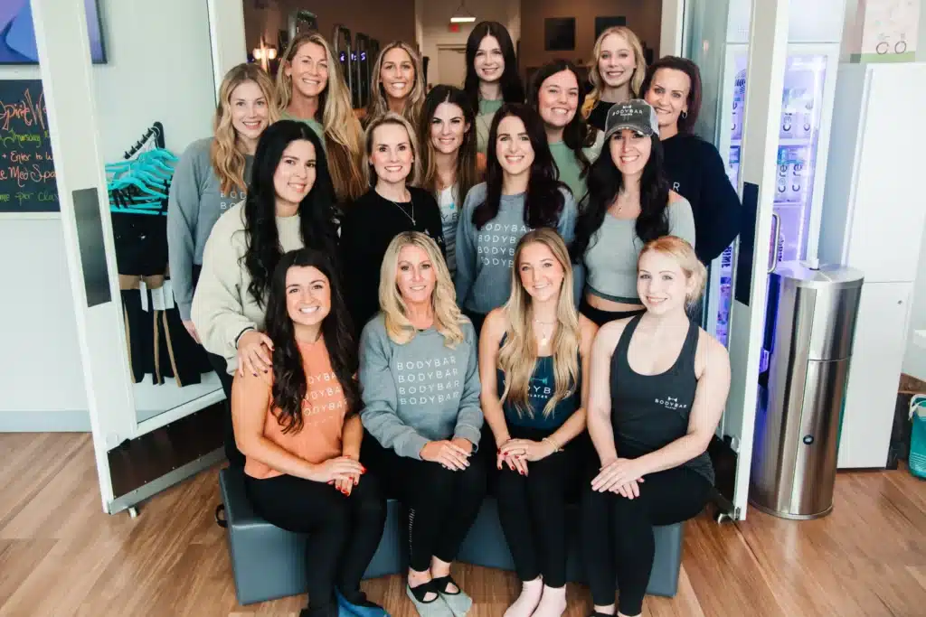 A group of sixteen women, smiling and posing together in a fitness studio.