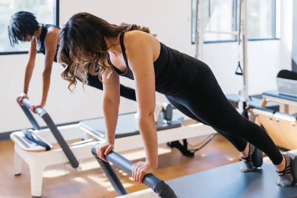 Two women in workout clothes use Pilates reformer machines in a bright fitness studio.