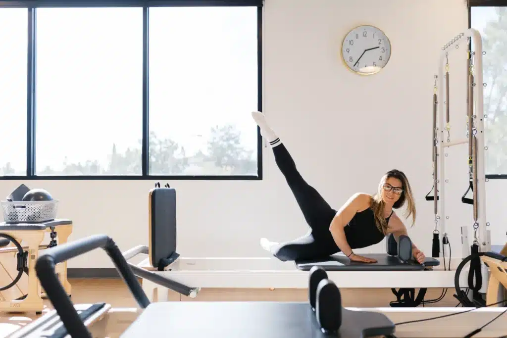 Woman in workout clothes does a side-lying leg lift on a Pilates reformer in a bright, modern studio.