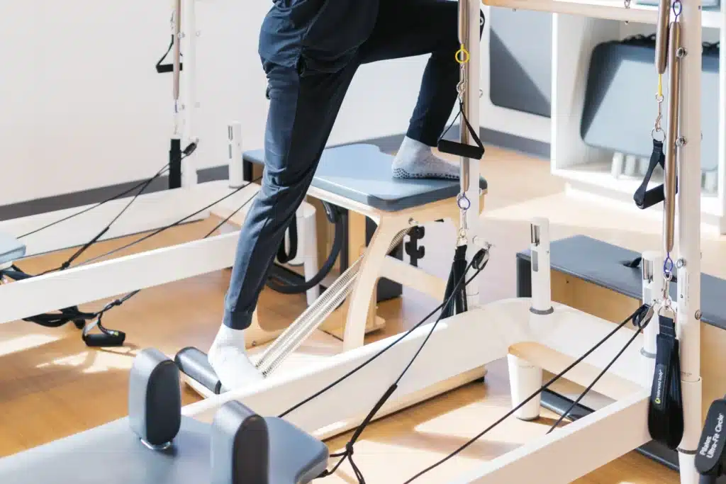 Person in black workout clothes using a Pilates reformer machine in a bright studio.
