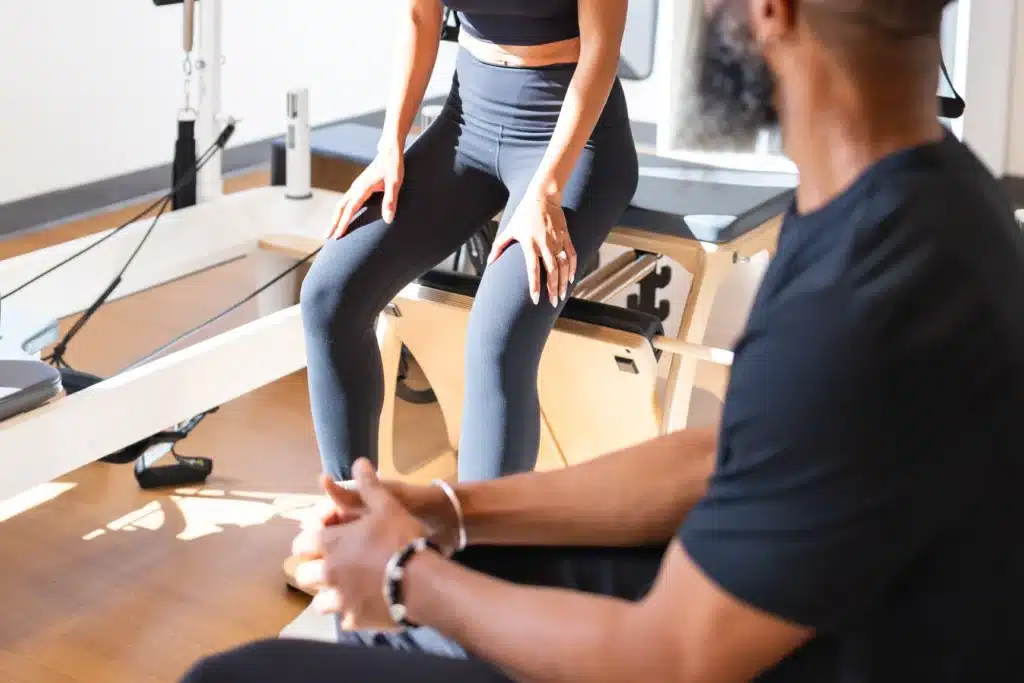 Two people in athletic wear sit in a bright gym, one on a reformer machine, the other facing them, talking.