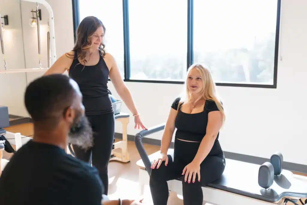 Two women in workout clothes talk as one sits on exercise equipment; a man listens in the foreground.