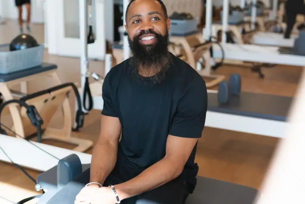 Man with a beard wearing a black shirt sits smiling on gym equipment in a bright fitness studio.