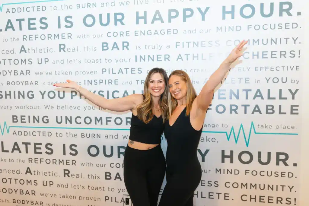 Two women in black workout clothes smile and pose with arms raised in front of a motivational fitness-themed wall.