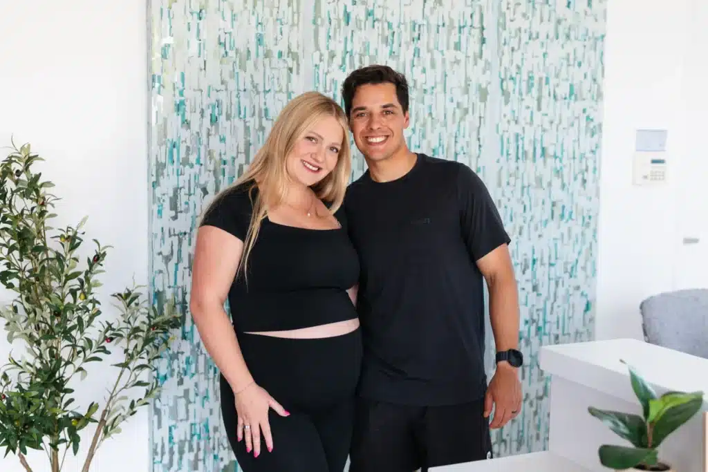Smiling couple stands indoors; the woman is pregnant and both wear black outfits. Plants and patterned curtains are visible.