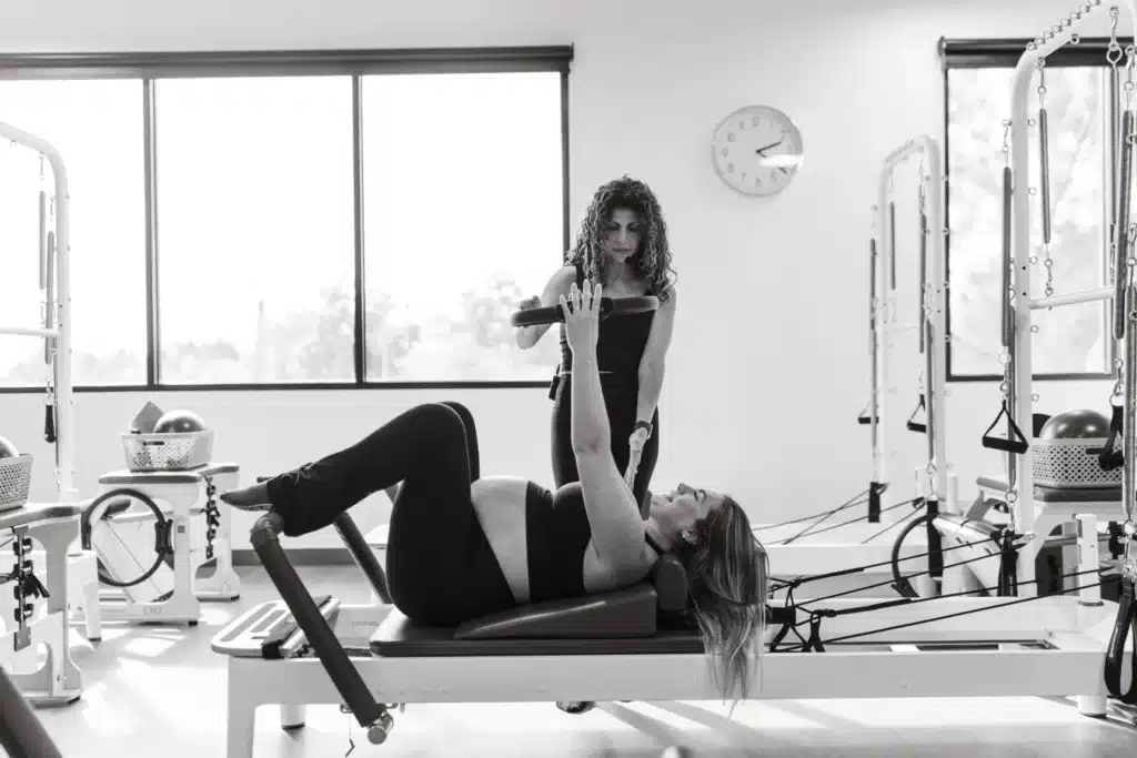 A woman works out on Pilates equipment while an instructor stands nearby in a bright, modern studio.
