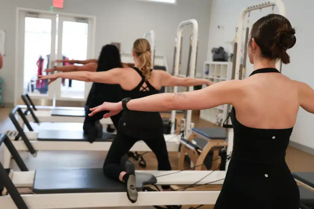 Three women in workout attire do Pilates on reformer machines in a bright fitness studio, arms extended for balance.