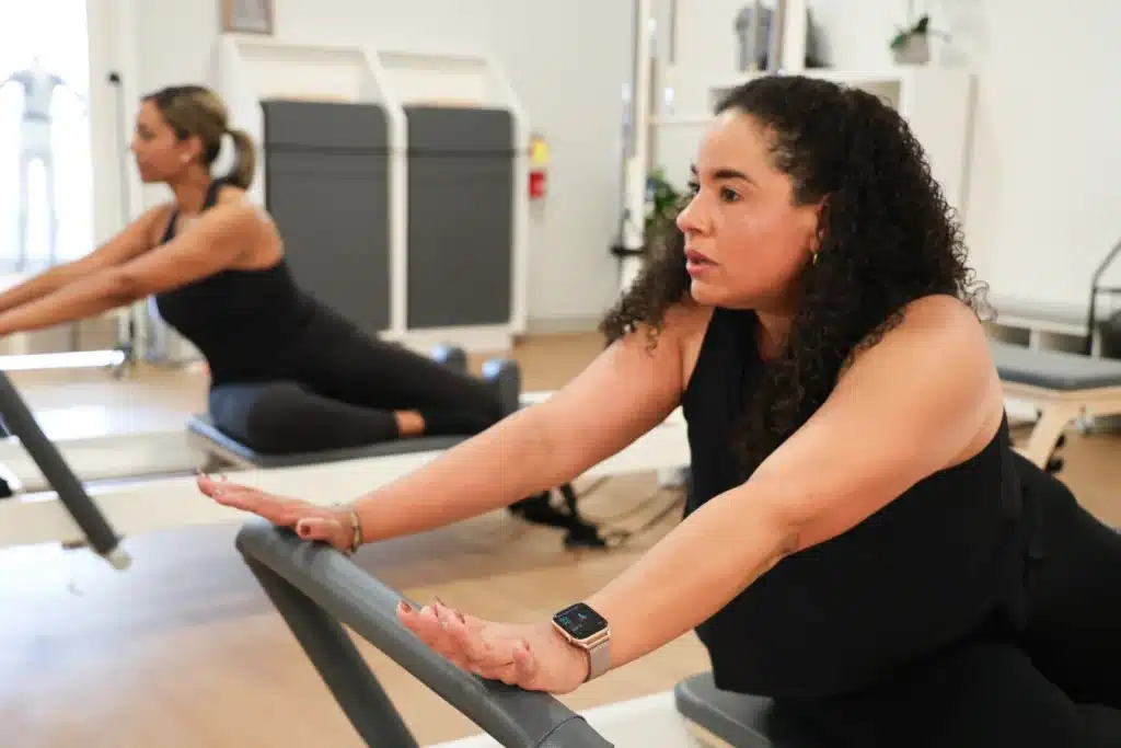 Two women in black workout clothes stretch on Pilates reformer machines in a bright, modern studio.