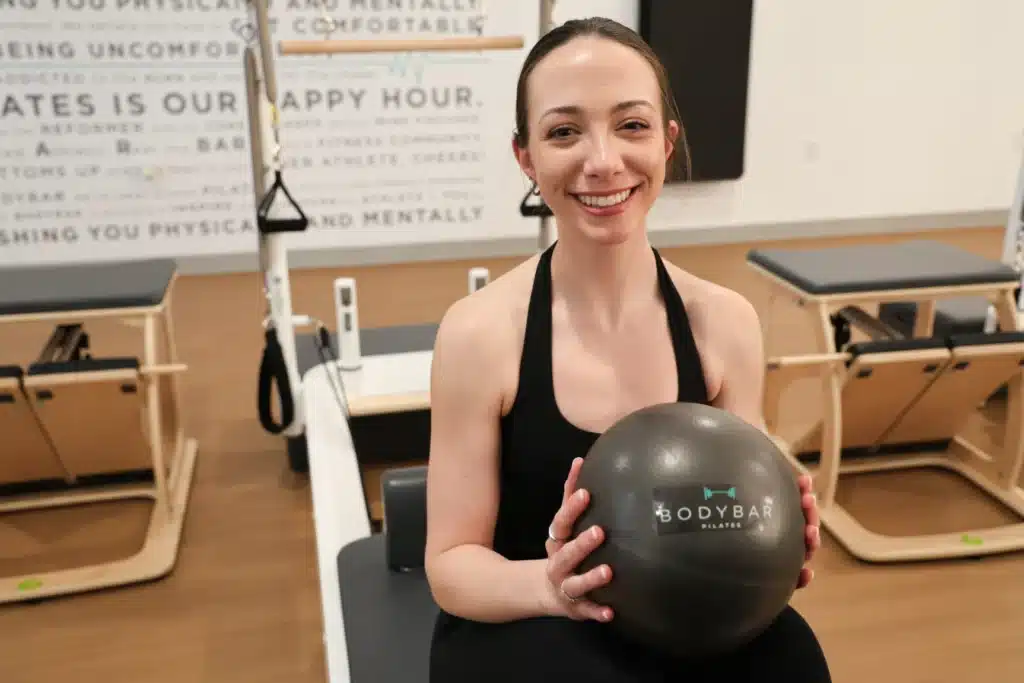 Woman smiling and holding a Pilates ball in a fitness studio with reformer machines in the background.