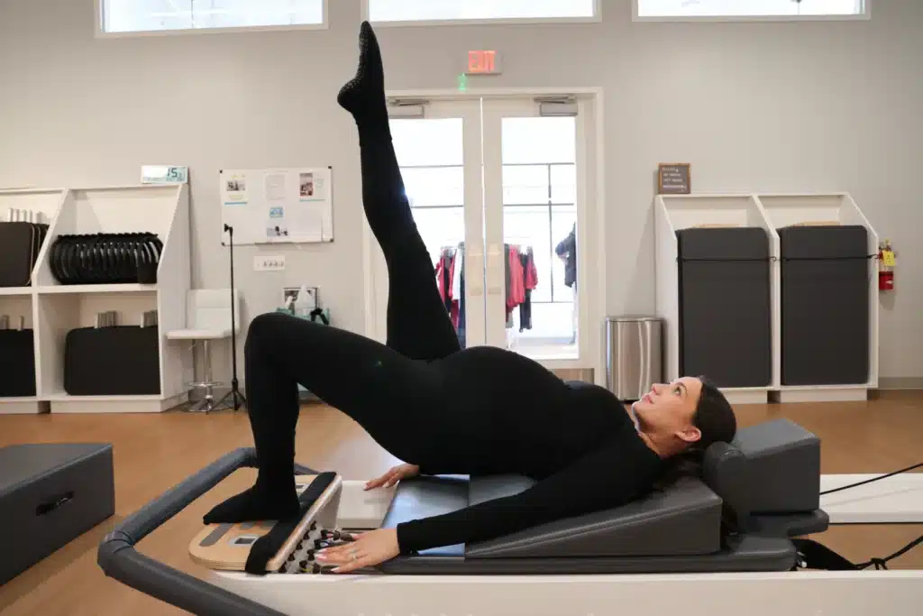 Pregnant woman in black workout clothes does Pilates on a reformer, raising one leg while lying on her back in a bright studio.