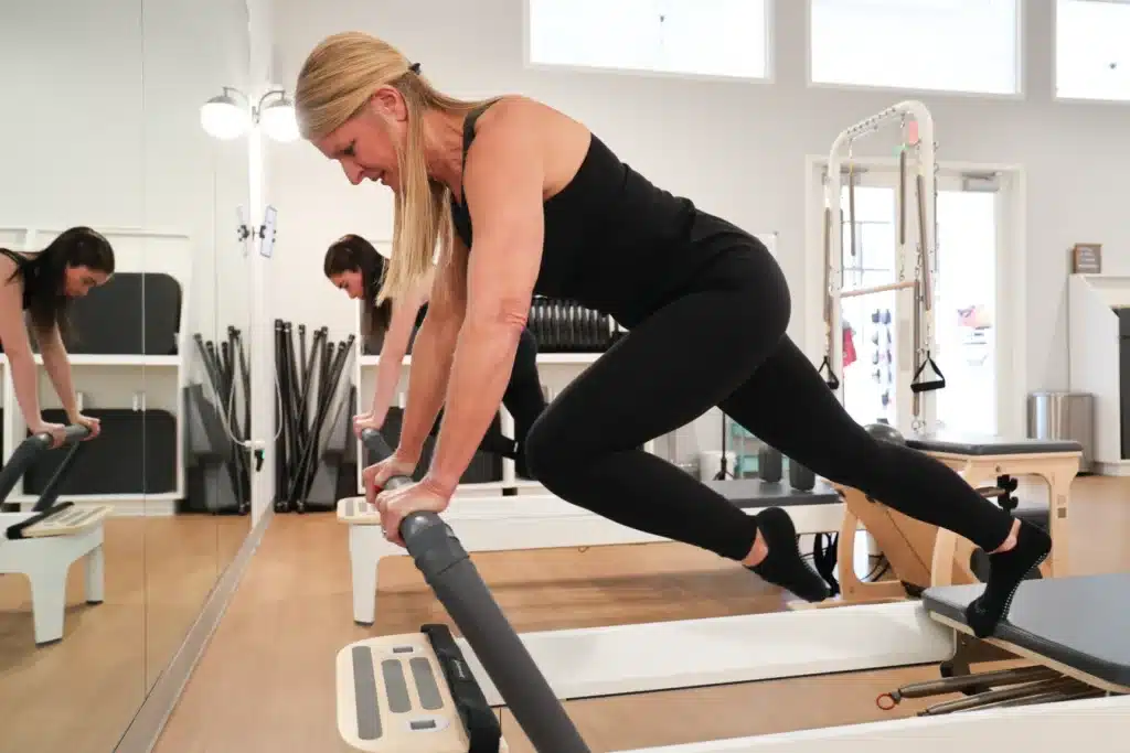 Two women exercise on Pilates reformer machines in a bright studio, both wearing black workout clothes.